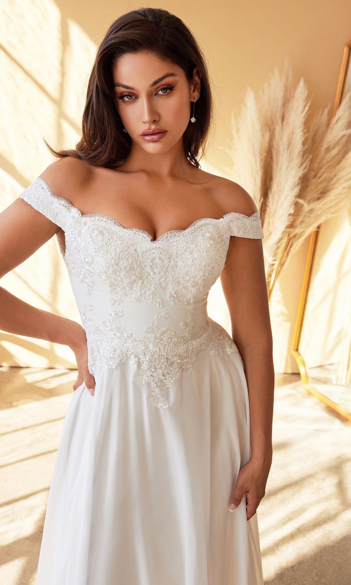 A woman stands indoors in warm lighting among neutral pampas grass, wearing the elegant and timeless Long Off-White Wedding Dress: CL7258W adorned with white lace—an affordable off-the-shoulder bridal gown.